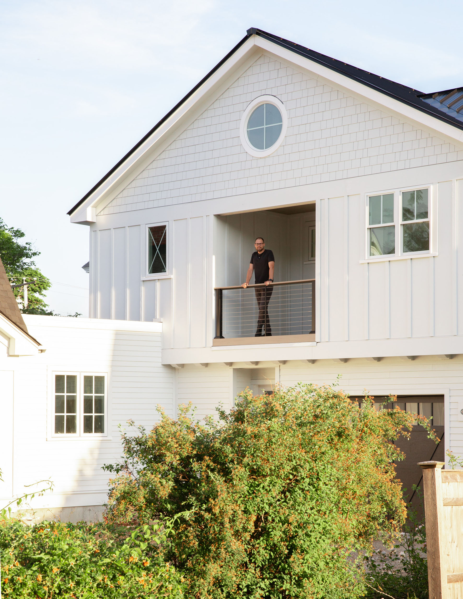 Man standing on a second-floor recessed balcony with cable railing on a modern white board-and-batten home featuring a round gable window and cedar shake accents